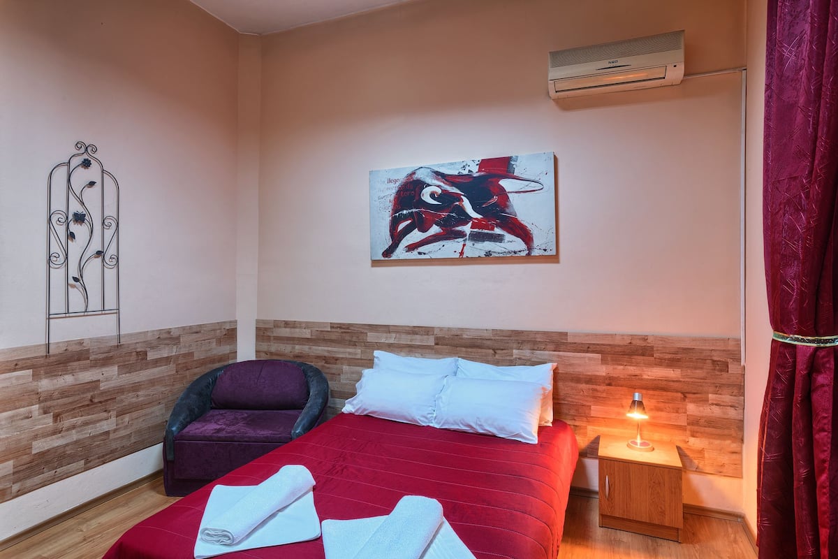 A hotel room with a wooden headboard, a bed with red bedding, a purple chair, a wall-mounted air conditioner, and a painting of a bullfight.