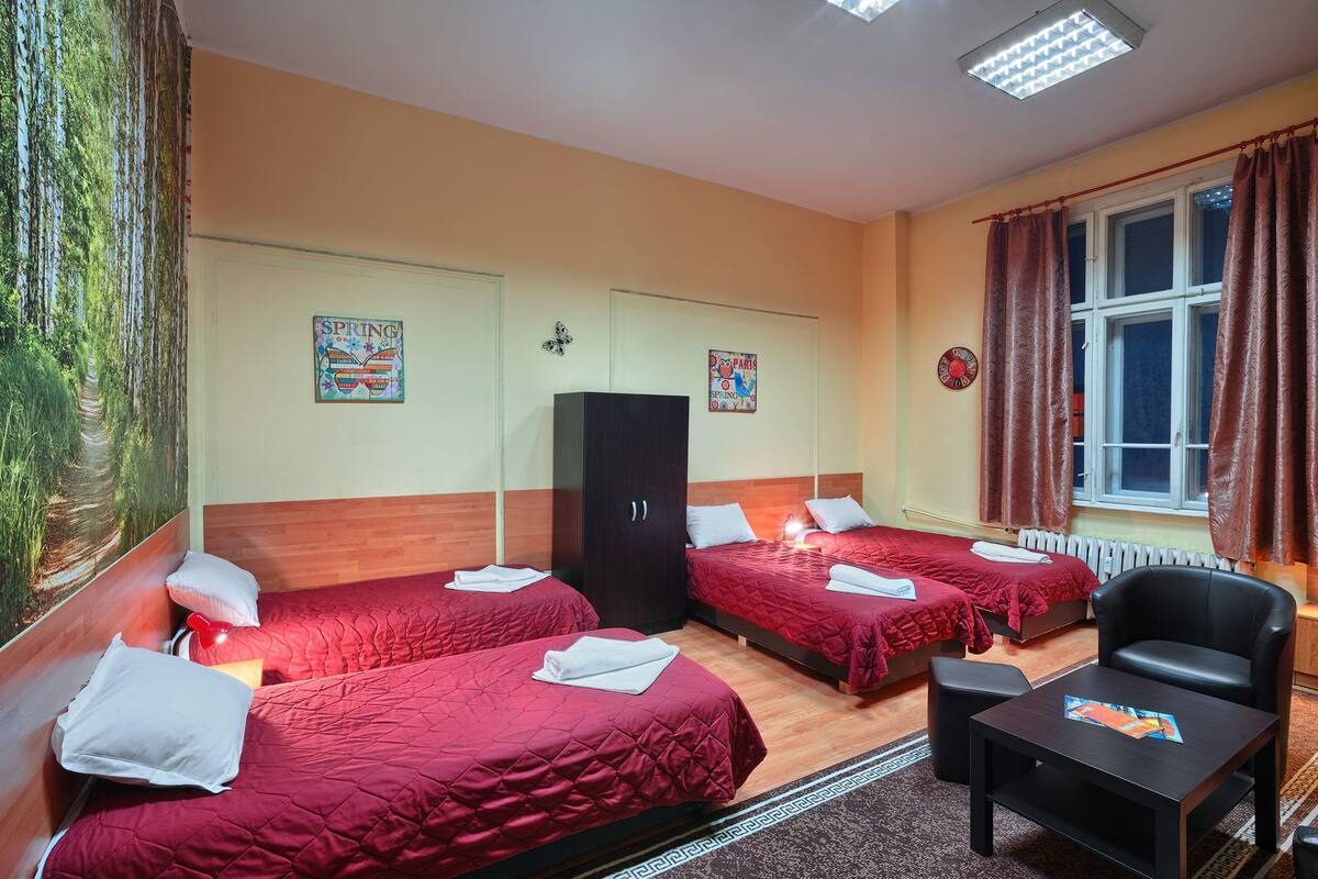 A hotel room with three beds, a wooden wardrobe, a desk with a chair, and a window with curtains.