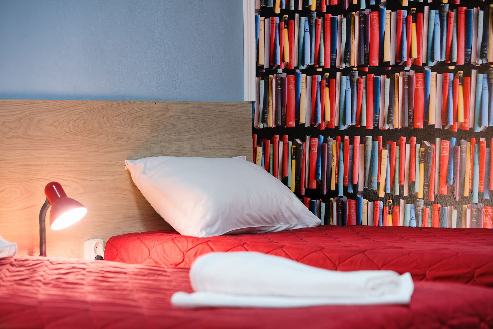 Un lit bien fait, recouvert d’une couverture rouge, avec un oreiller blanc et une serviette blanche roulée. Une lampe rouge est posée sur la table de chevet, et une bibliothèque remplie de livres se trouve à l’arrière-plan.