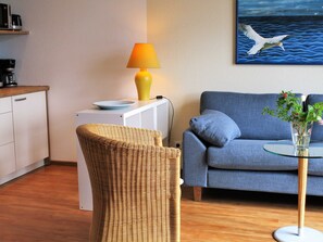 A room with a wicker chair, a blue sofa, a glass table with a vase, a lamp, and a painting of seagulls.