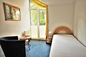 Une chambre d’hôtel avec un lit, une chaise, une petite table, une lampe et une fenêtre avec des rideaux.