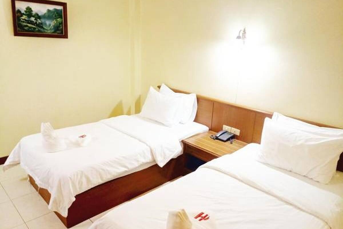 Quarto de hotel com duas camas de solteiro, roupa de cama branca, cabeceira de madeira, mesa de cabeceira com telefone e um quadro na parede.