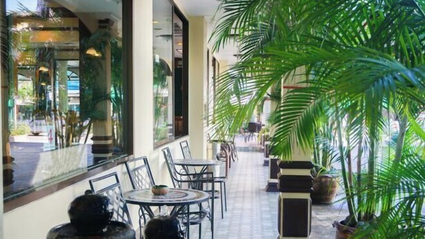 A covered outdoor seating area with tables and chairs, surrounded by large potted plants and a glass wall reflecting the outside scene. - Hua Lin Inn (Chiang Mai)