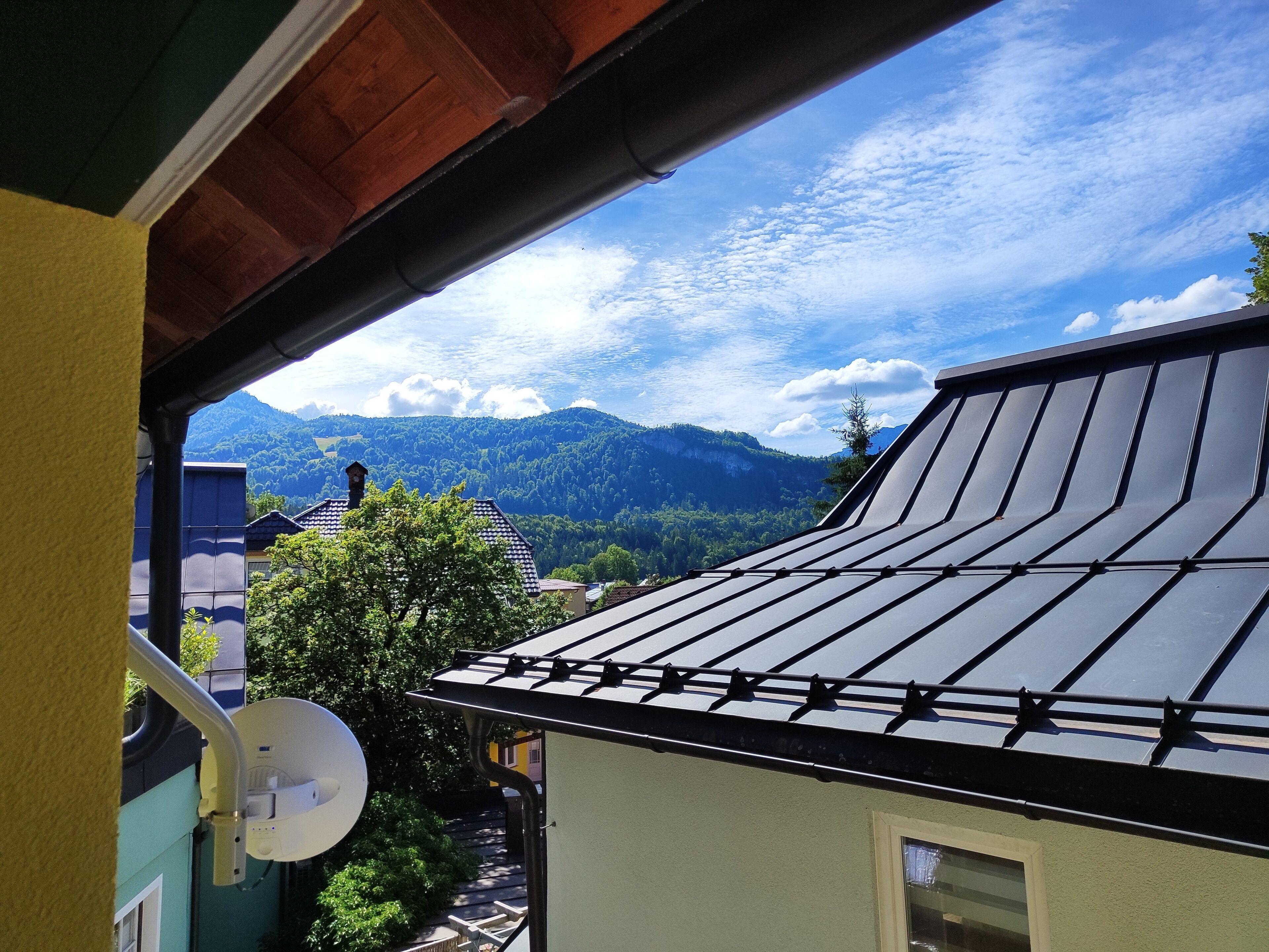 Une vue depuis un balcon, avec en arrière-plan une montagne, une antenne parabolique et un bâtiment doté d’un toit en pente.