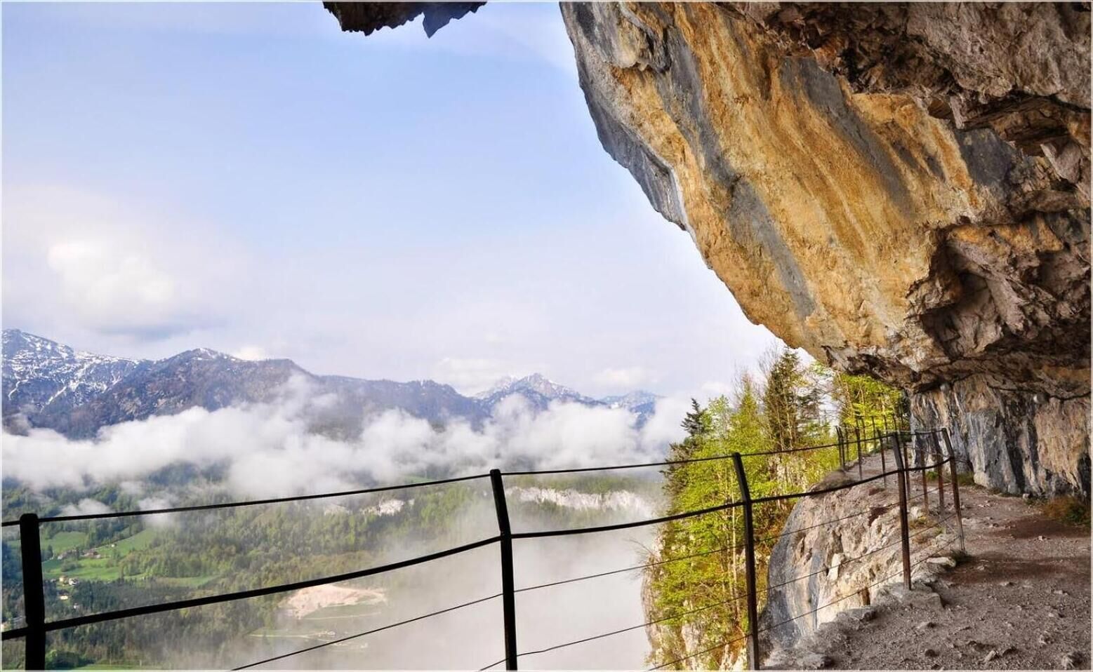 A mountainous landscape with a clear view of a valley and a river, viewed from a rocky ledge with a safety railing.