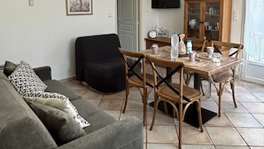 A living room with a grey sofa, a black armchair, a wooden dining table with chairs, a television mounted on the wall, and a cabinet with glass doors.