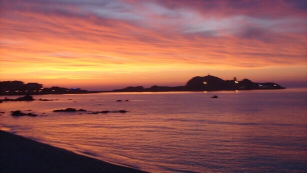 A beach at sunset with calm waters and distant land silhouettes.