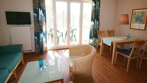 A room with a sofa, a dining table with chairs, a TV mounted on the wall, and a window with curtains.