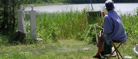 Uma pessoa pintando à beira de um lago, com uma cadeira e cavalete, rodeada por relva e árvores.