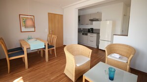 A room with a dining table, chairs, a small kitchen area, and wicker furniture.