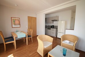 A room with a dining table, chairs, a small kitchen area, and wicker furniture.