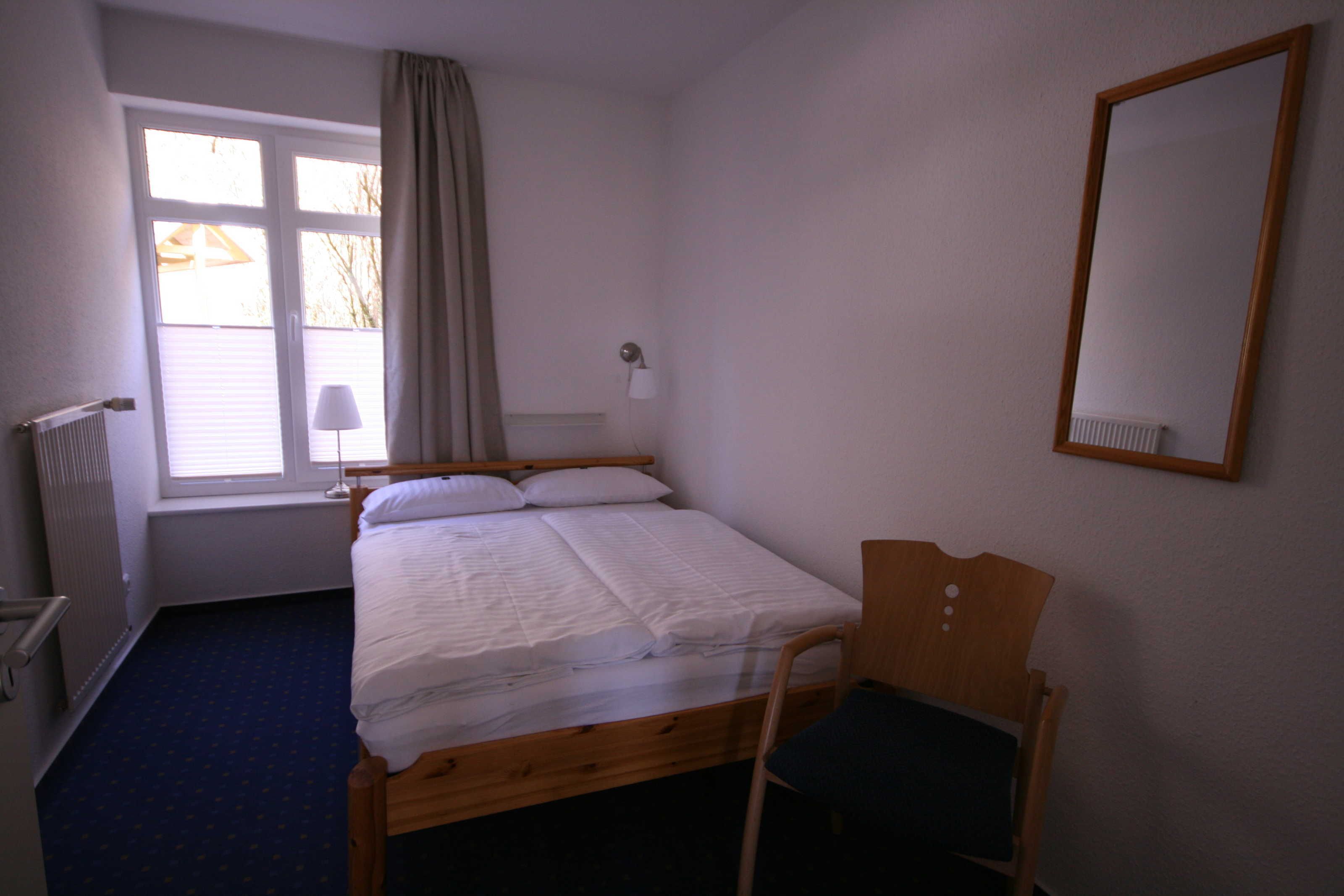 A single bed with a wooden frame, a chair, a window with curtains, a wall-mounted lamp, and a mirror.