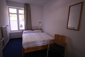 A single bed with a wooden frame, a chair, a window with curtains, a wall-mounted lamp, and a mirror.
