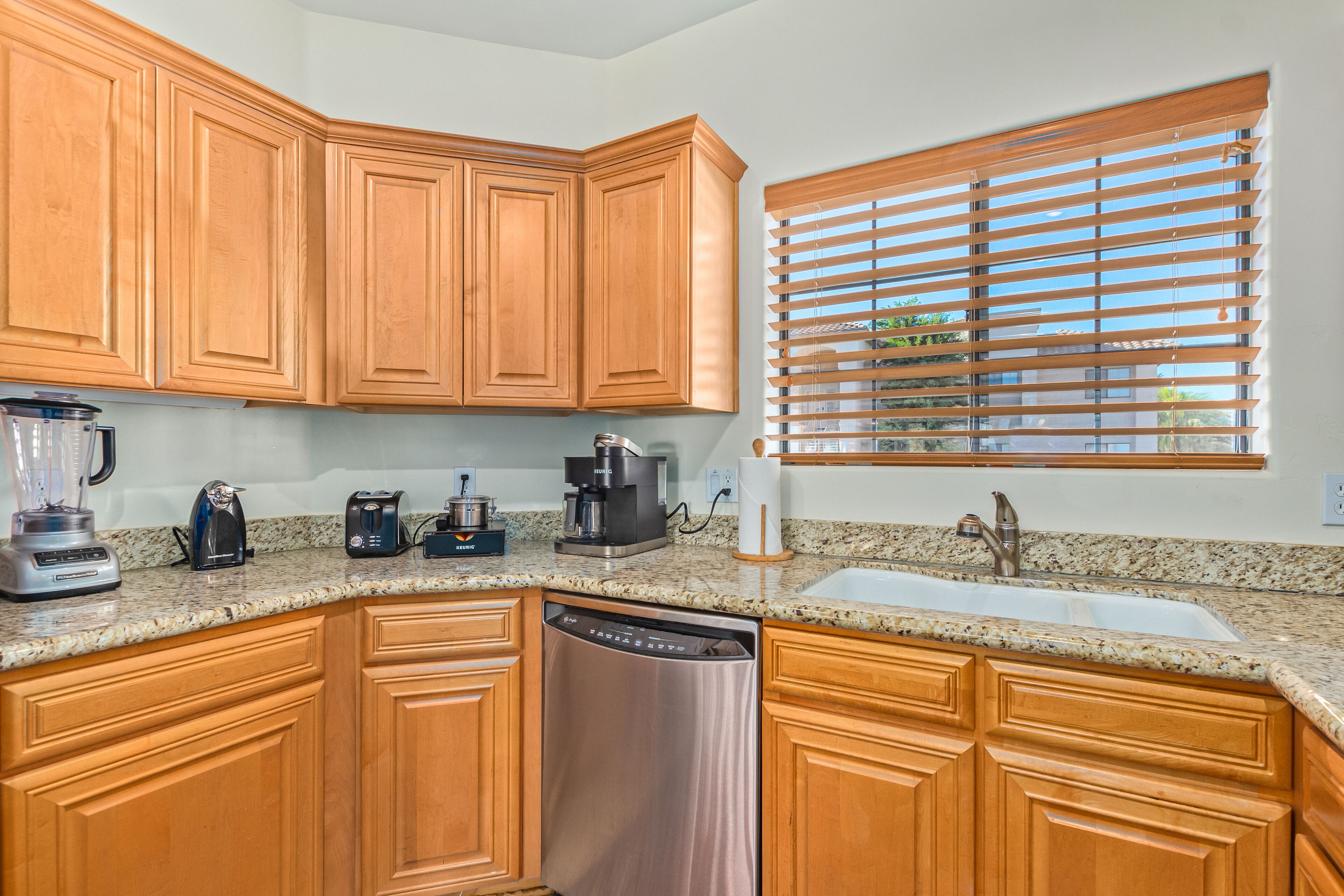 A kitchen with wooden cabinets, granite countertops, a dishwasher, a coffee maker, and a window with blinds.