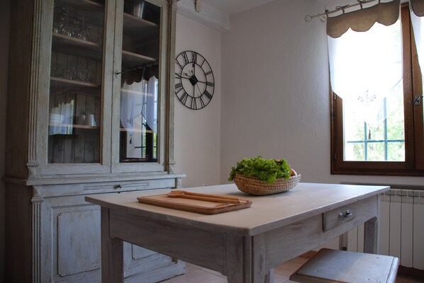 A kitchen with a wooden table, a clock, and a window with curtains. - description (Lorgues)
