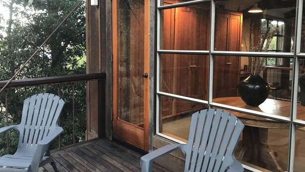 Een houten terras met twee Adirondack-stoelen, een glazen deur naar een kamer met houten kasten en een vaas op een tafel.