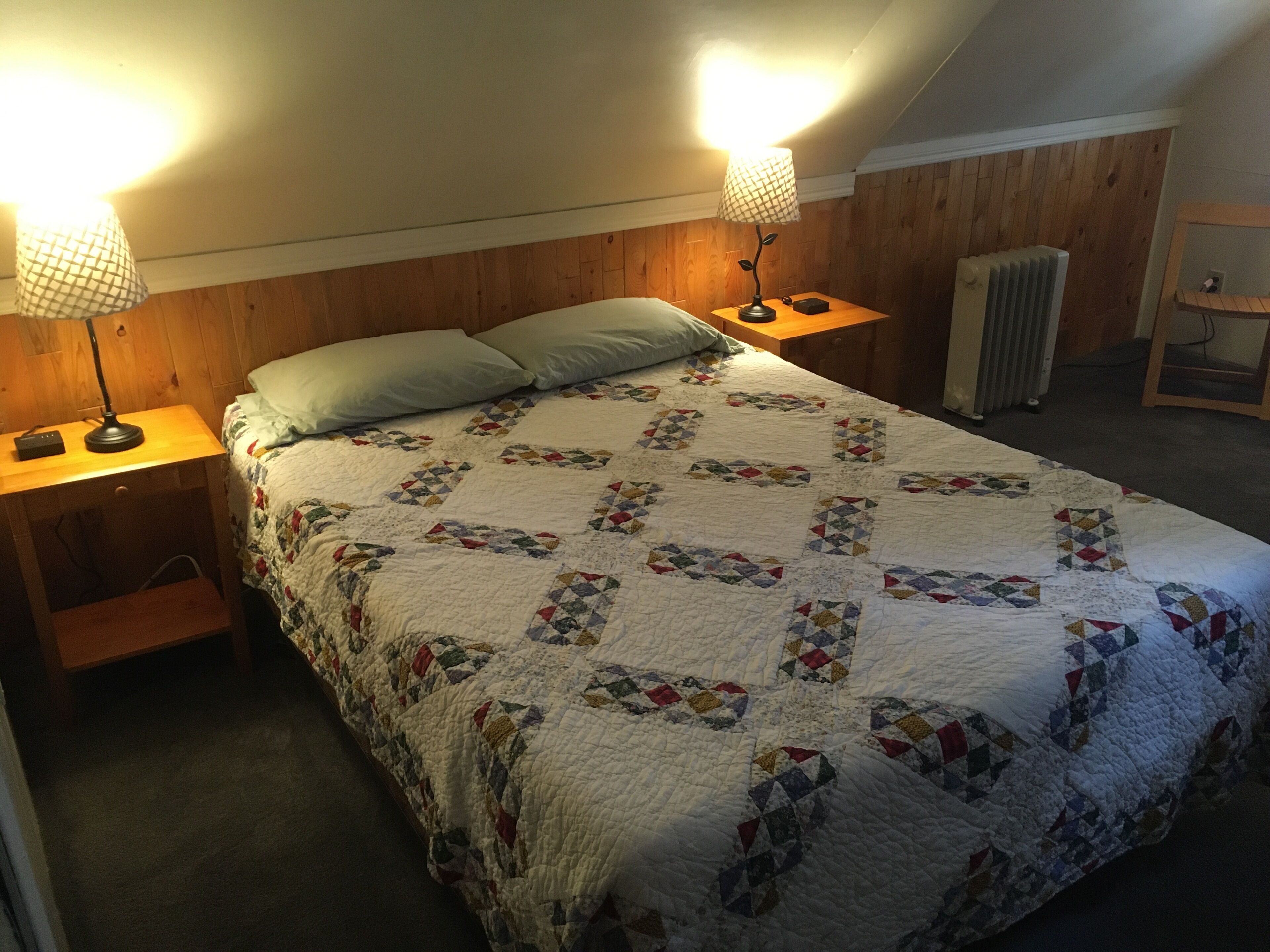 Un dormitorio con una colcha acolchada, dos lámparas de noche, un cabecero de madera y un radiador.