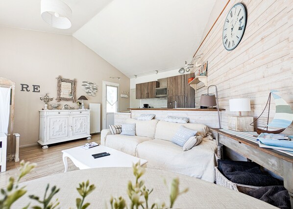 A cozy living room with a white sofa, a coffee table, and a bookshelf. There is a kitchen area with wooden cabinets and a microwave. A large clock is visible on the wall.
