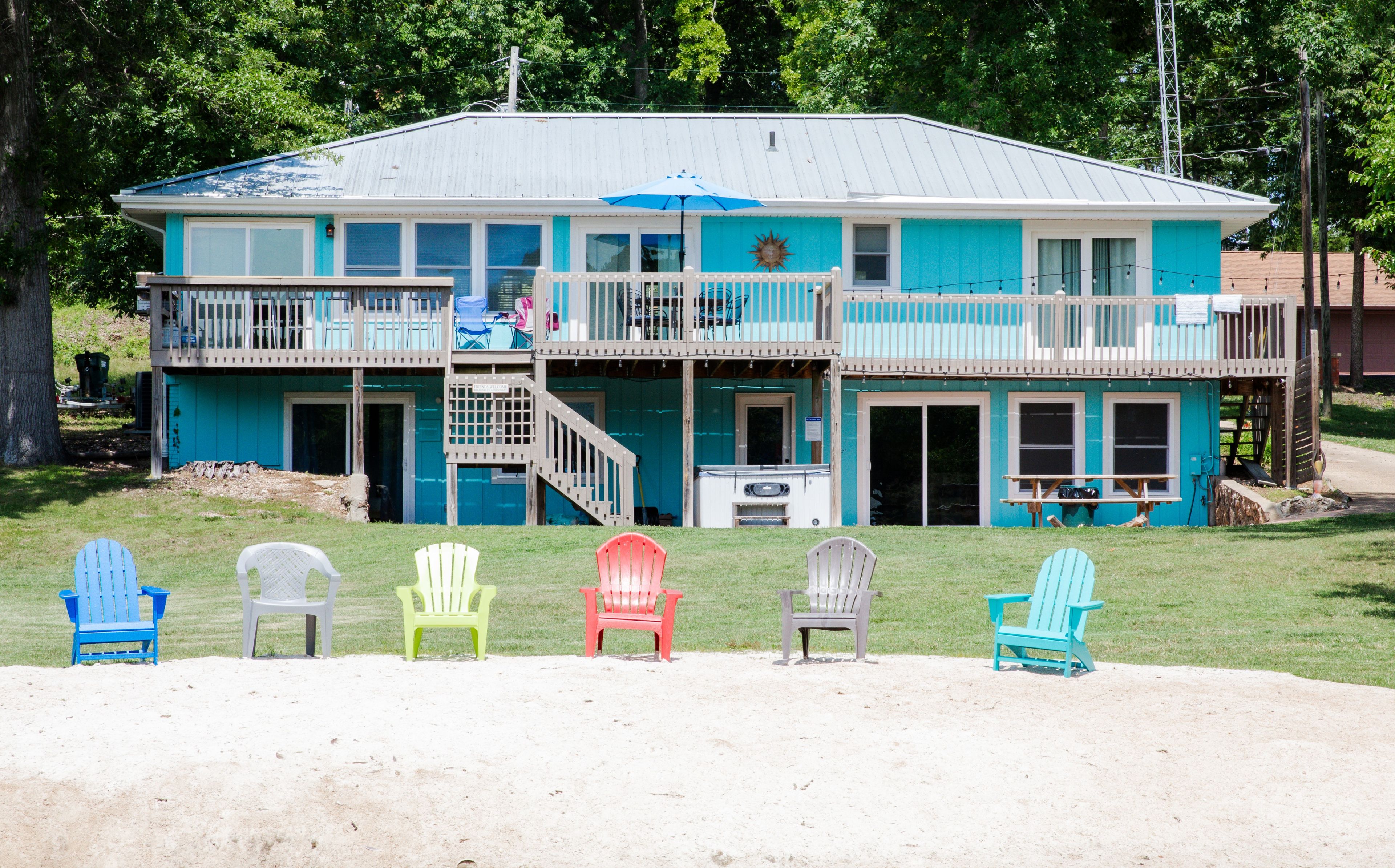 A two-story building with a balcony, a covered patio, and a sandy area with Adirondack chairs.