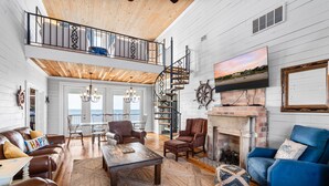 A spacious living room with a high ceiling, a wooden staircase, a fireplace, and a balcony with a view. - Beachfront Property in Ocean Springs, Overlooking the Gulf of Mexico. Sleeps 14 (Ocean Springs)