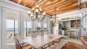A spacious dining area with a wooden table and chairs, a bench, and a view of the ocean through large windows. - Beachfront Property in Ocean Springs, Overlooking the Gulf of Mexico. Sleeps 14 (Ocean Springs)