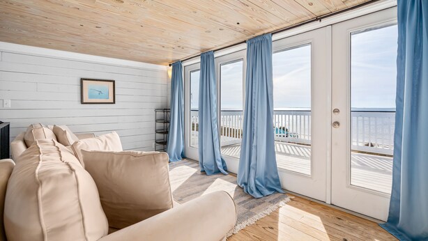 A room with a wooden ceiling, a beige sofa, a framed picture on the wall, and large windows with blue curtains. - Beachfront Property in Ocean Springs, Overlooking the Gulf of Mexico. Sleeps 14 (Ocean Springs)