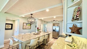 A modern kitchen with a marble island, white bar stools, a stainless steel refrigerator, and decorative items on shelves. - Beautifully Updated Ground Floor Condo, Direct OCEAN Front on NO-DRIVE Beach! (New Smyrna Beach)