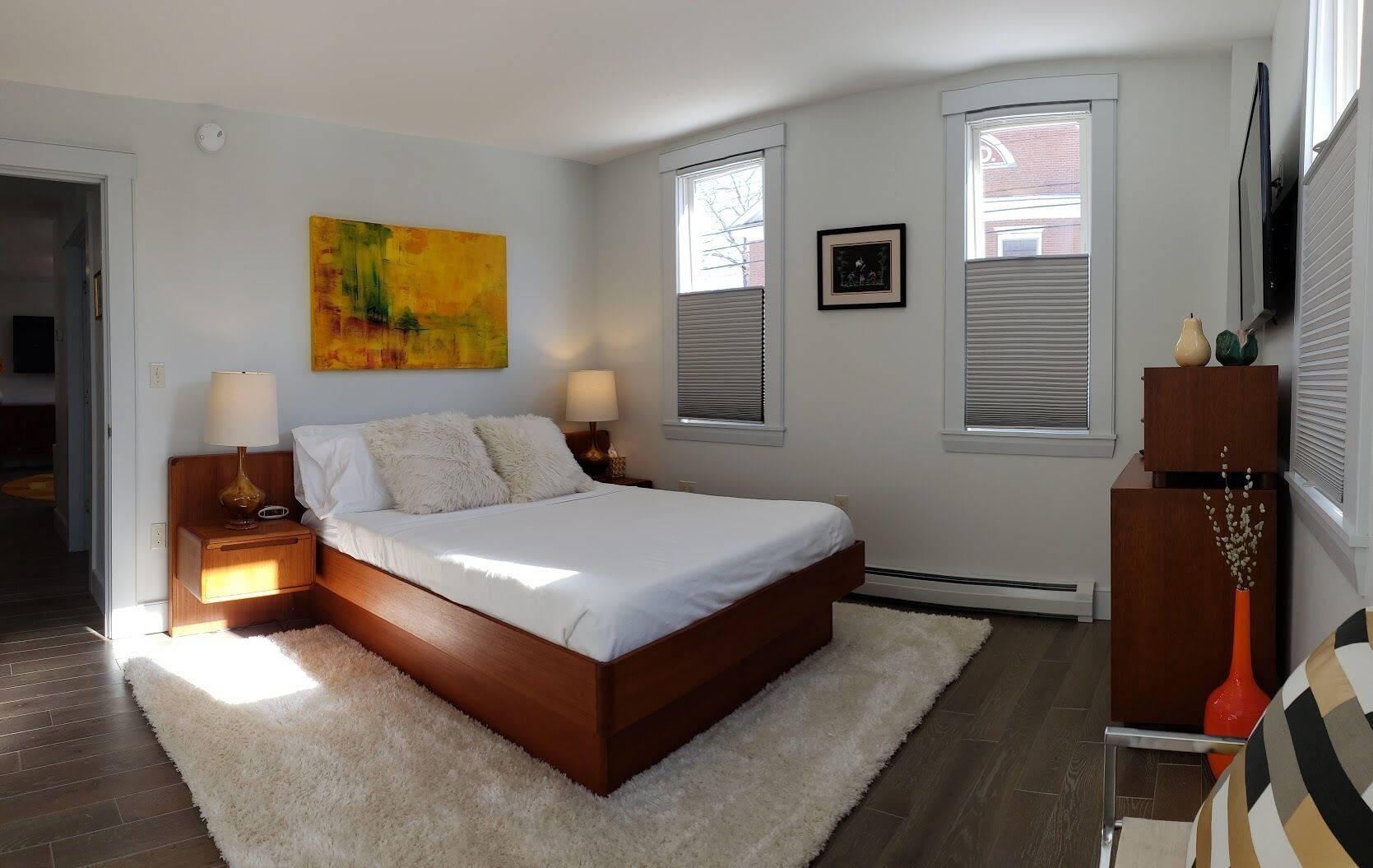 Quarto com cama de madeira, roupa de cama branca, duas luminárias de cabeceira, tapete macio, um quadro, uma fotografia emoldurada e um penteado com objetos decorativos.