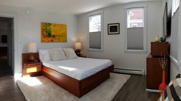 Quarto com cama de madeira, roupa de cama branca, duas luminárias de cabeceira, tapete macio, um quadro, uma fotografia emoldurada e um penteado com objetos decorativos.
