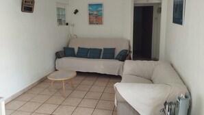 Uma sala de estar com um sofá bege, uma mesa de madeira redonda e uma poltrona branca. Há um quadro na parede e uma porta que dá para outro cômodo.