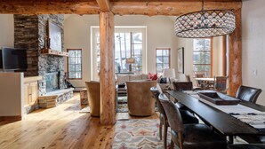 A spacious living area with a stone fireplace, wooden beams, a large dining table, and a chandelier. - Harvest Moon Haven | Premier Ski Access + Hot Tub (Big Sky)