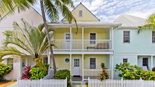 A two-story house with a front porch, a white picket fence, and tropical plants.