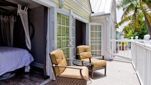 A balcony with two chairs and a table, a bed, and a sliding glass door leading to a room.