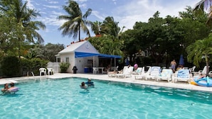 A swimming pool with clear water, surrounded by lounge chairs and umbrellas, with a small building and palm trees in the background.
