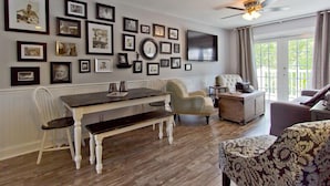 A dining area with a wooden table and chairs, a television mounted on the wall, and a ceiling fan.