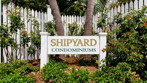 A sign for Shipyard Condominiums surrounded by lush greenery and a white picket fence.