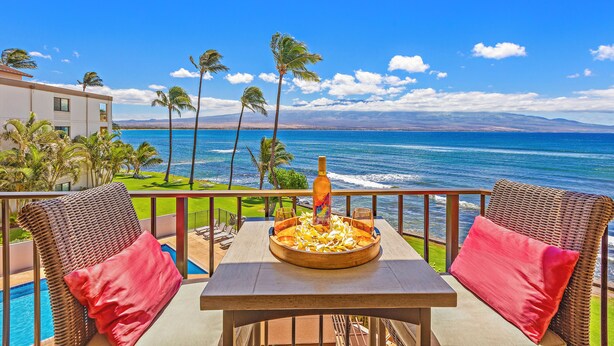 A balcony with wicker furniture, a table with a bottle and glasses, and a view of the ocean and palm trees. - Great Listing/Price/AC in the bedroom! (Maalaea)