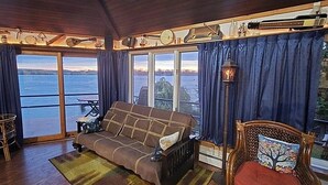 A cozy living room with a sofa, a wooden chair, and a view of the water through large windows.