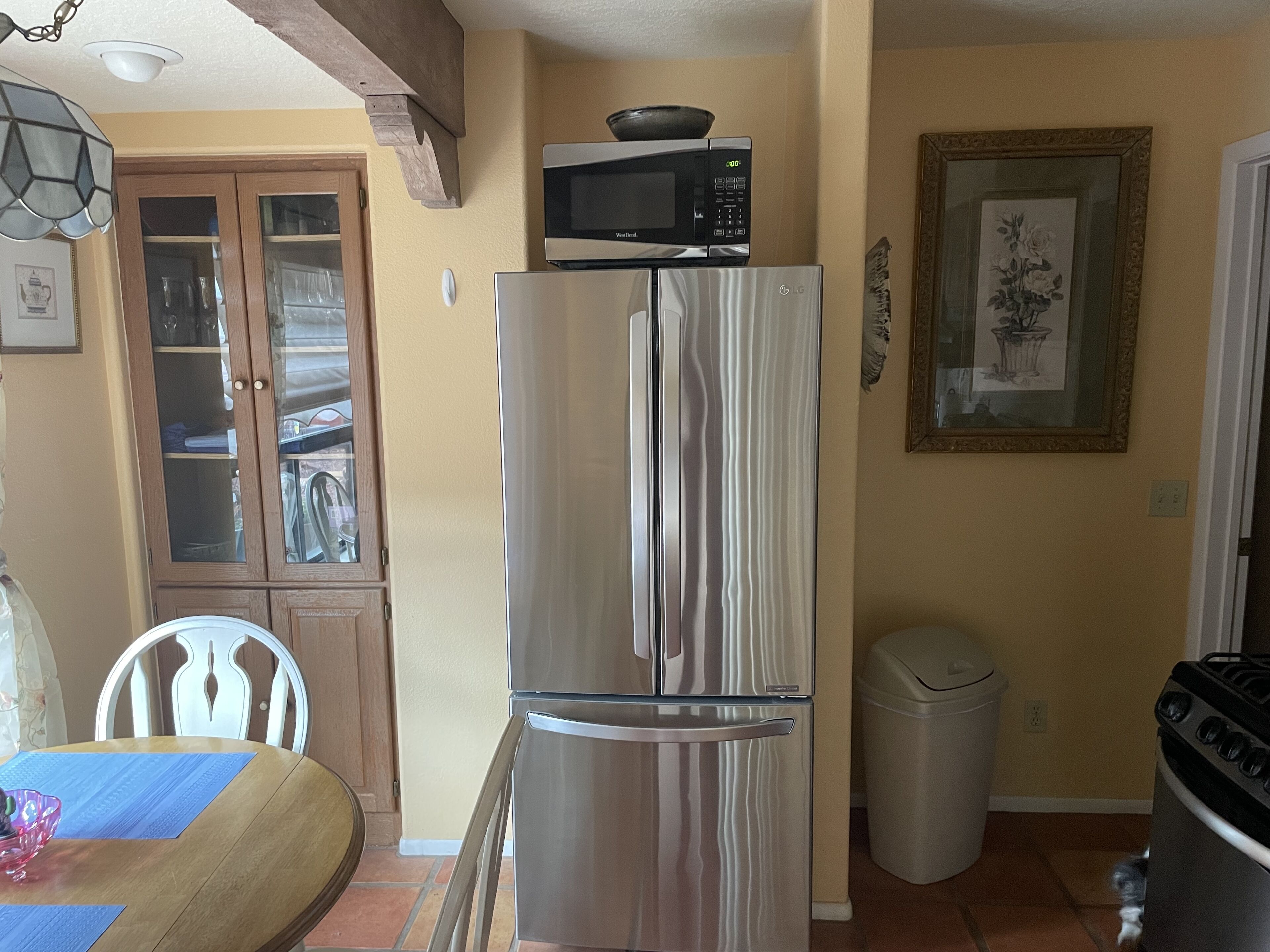 A kitchen with a stainless steel refrigerator, a microwave on top, a trash bin, and a framed picture on the wall.