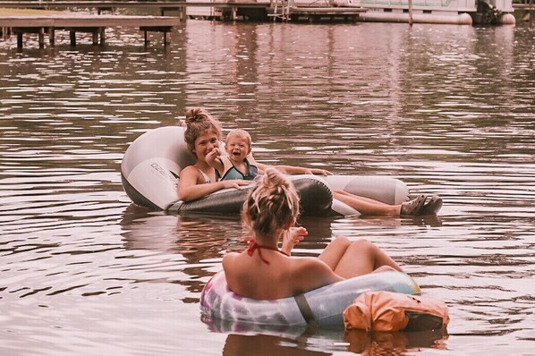 En grupp människor som flyter på en flotte på ett vattenområde, med en brygga och båtar i bakgrunden.