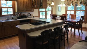 A kitchen with wooden cabinets, a central island with bar stools, a dining table with chairs, and large windows with curtains. - Beautiful 5 bedroom, 3.5 bath home on a quiet cove on Lake Gaston (Littleton)