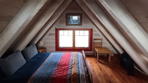 Un dormitorio acogedor en el ático, con techo inclinado de madera, una cama con manta colorida, un ventanal pequeño y una silla.