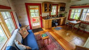 Interior acogedor de una cabaña con una mesa de comedor de madera, un sofá azul y una zona de cocina con armarios de madera y una ventana con marco rojo.