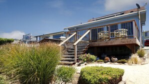 A coastal house with a wooden deck, a staircase leading to a balcony, and a sandy garden area.