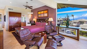 A spacious bedroom with a large bed, multiple seating options, a ceiling fan, and a view of a residential area.
