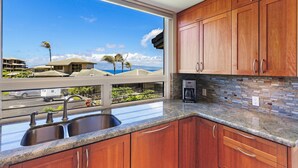A kitchen with wooden cabinets, a granite countertop, a large window with a view of a residential area and palm trees, and a coffee maker.