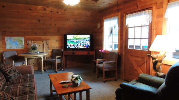 A cozy cabin-style room with wooden walls, a television, a dining table, and two sofas.