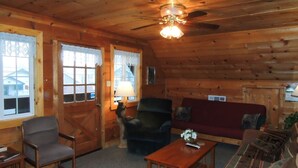 Interior acollidor d'una cabana de fusta amb sostre de fusta, un ventilador de sostre, un sofà vermell, un armari fosc, una taula de centre de fusta i una finestra amb cortines de gallet.