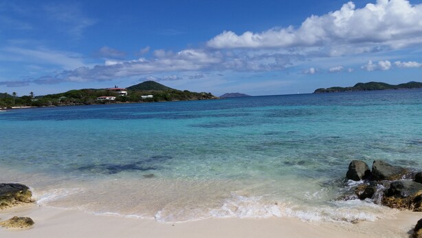 A beach with clear turquoise water, a sandy shore, and a small island with buildings in the background. - The Captain and the Mermaid - Adventures in Paradise are waiting! (St Thomas)
