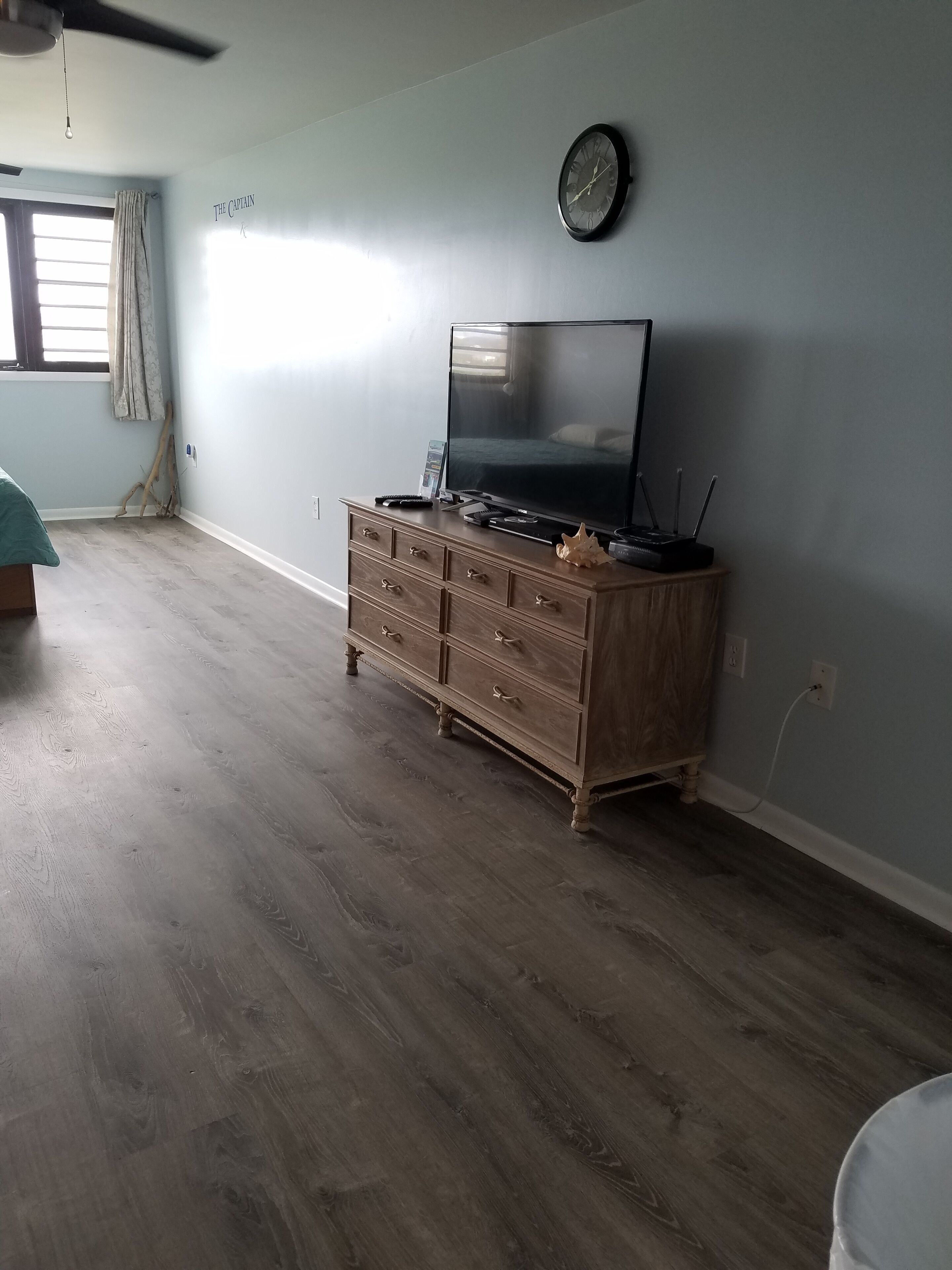A room with a wooden dresser, a flat-screen TV, a clock on the wall, and a window with curtains.